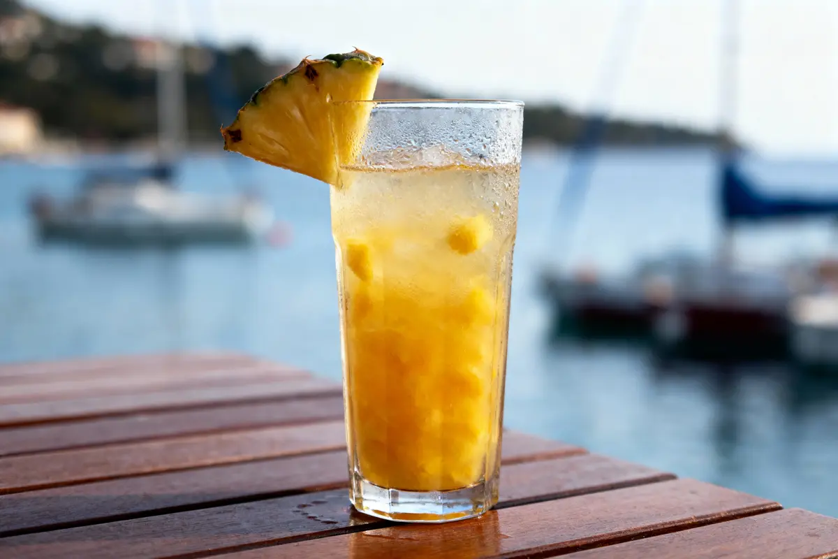 pexels-michal-pech-213601-757318 (1) Verre de cocktail à l'ananas, table en bois, vue sur la mer et bateaux en arrière-plan.