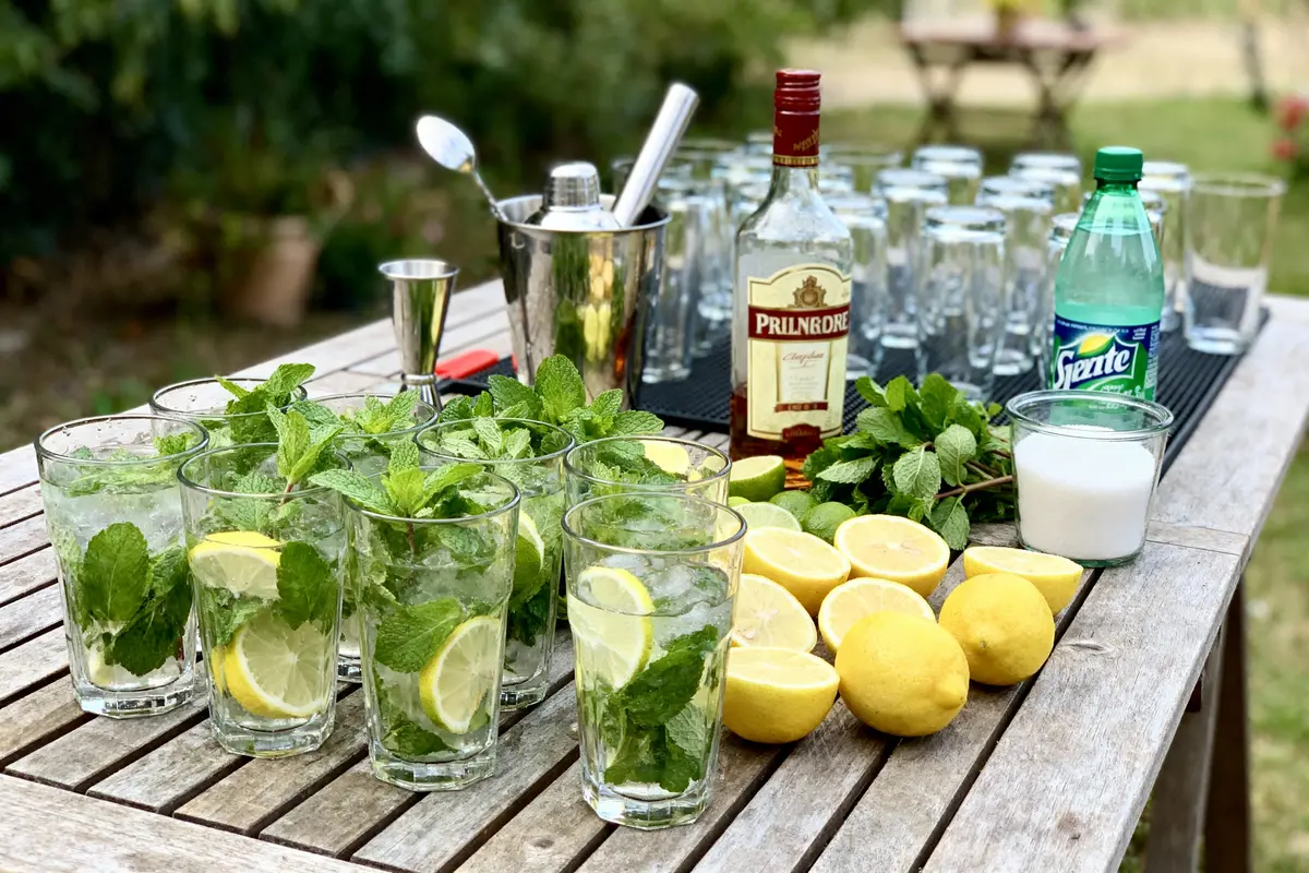 Verres de mojito préparés sur table bois avec citrons, menthe, bouteille de rhum, sucre, eau gazeuse en extérieur.