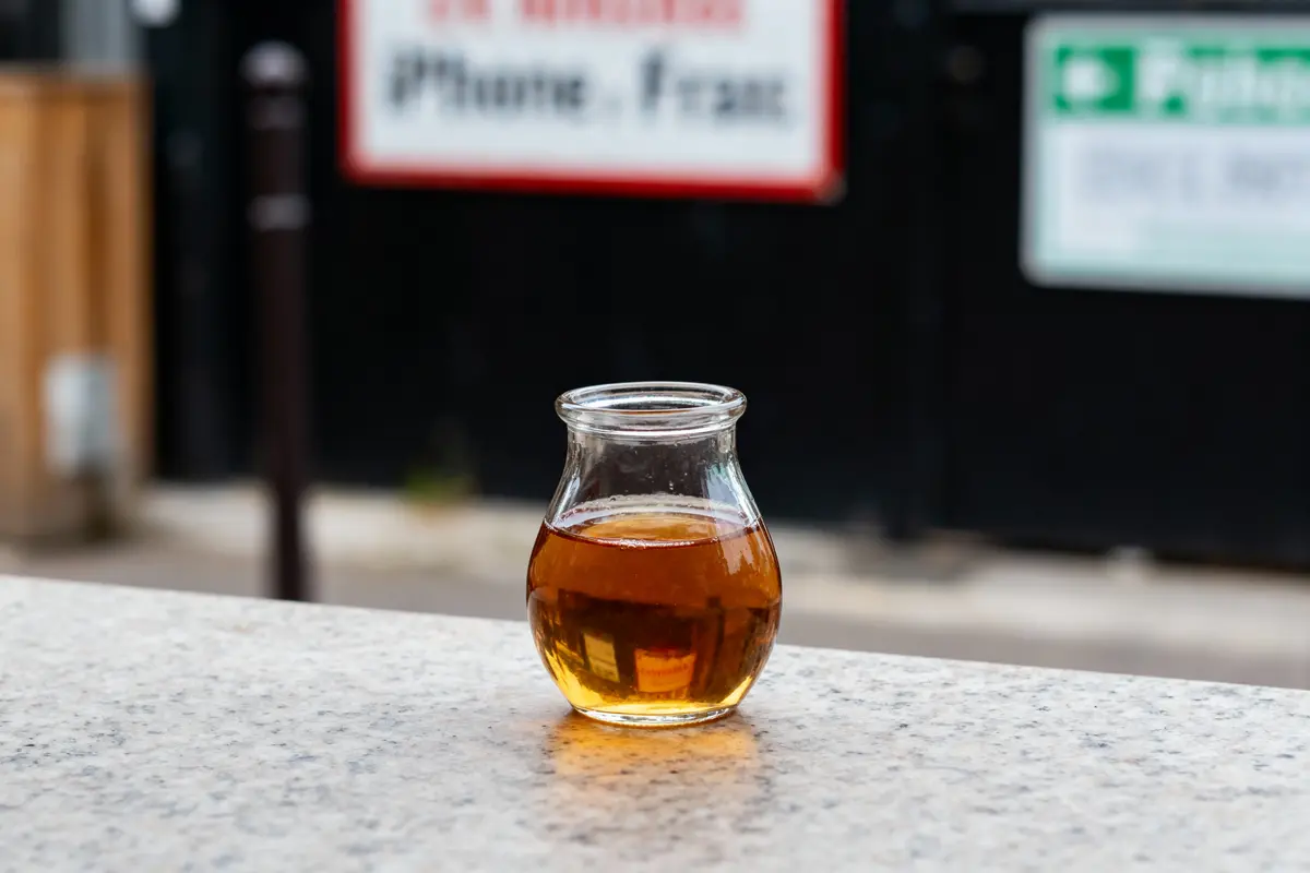 Un petit pot en verre avec liquide ambré sur une table en extérieur, arrière-plan flou.
