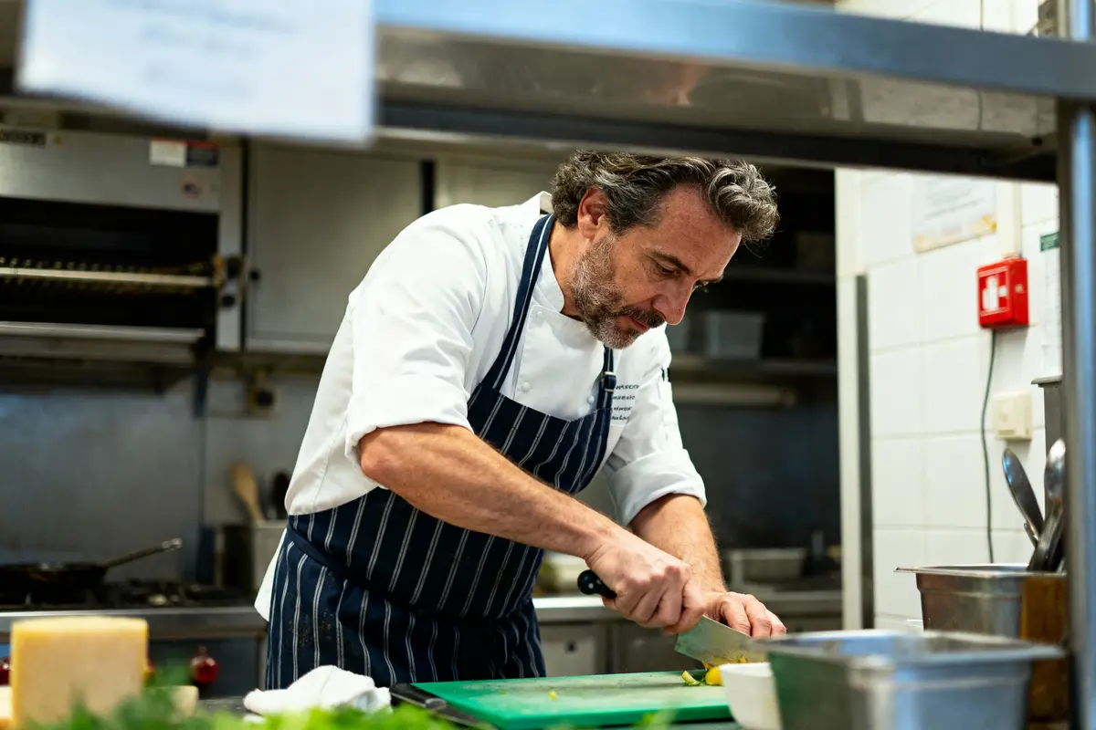 pexels-michal-pech-213601-757318 (1) Chef en veste blanche et tablier rayé coupant des légumes dans une cuisine professionnelle.
