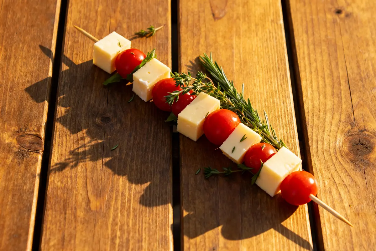 Brochette avec cubes de fromage, tomates cerises, herbes fraîches, sur table en bois sous lumière ensoleillée.
