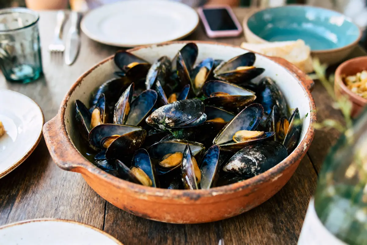 Plat de moules cuites dans un bol en terre cuite sur une table en bois, entouré d'assiettes et verres.