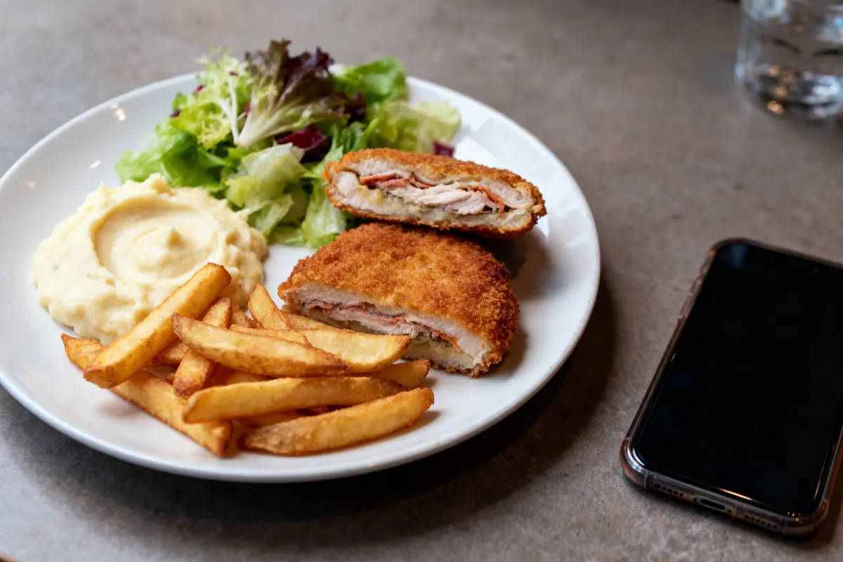 Assiette avec cordon bleu, frites, purée, salade verte. Téléphone noir sur la table.
