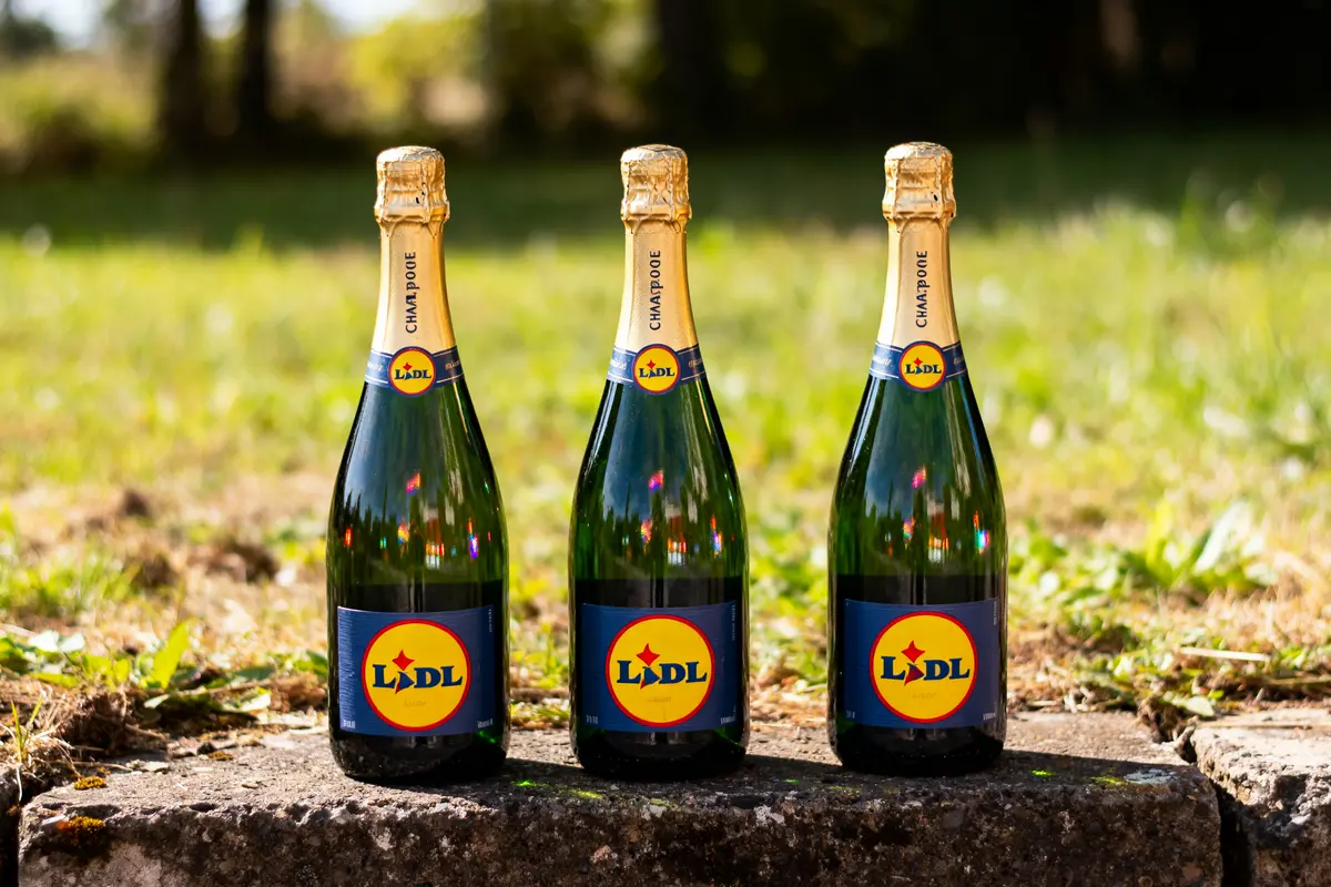 Trois bouteilles de champagne marquées Lidl posées sur un muret en extérieur, entourées de verdure.